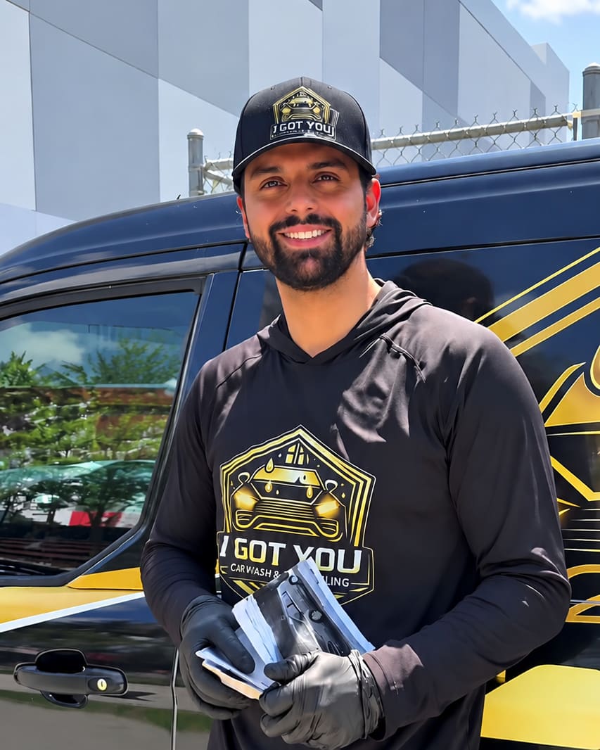 A smiling person in an I Got You cap and hoodie, wearing work gloves and holding supplies, standing beside the branded van.